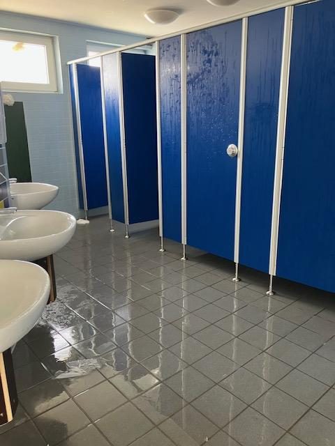 Toilettenraum mit blauen Kabinentüren, Wasser auf dem Boden, Waschbecken links.