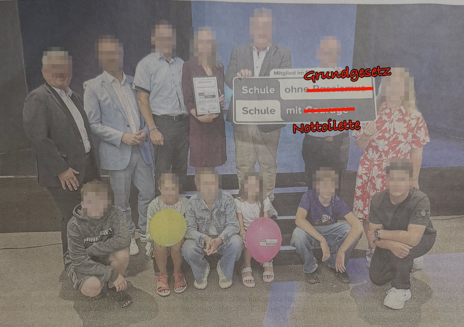 Satirisch bearbeitetes Foto der Uebergabe: Erwachsene und Kinder auf einer Buehne, alle Gesichter unkenntlich gemacht. Auf dem Schild sind die Woerter Rassismus und Courage rot durchgestrichen und durch Grundgesetz und Nottoilette ersetzt.