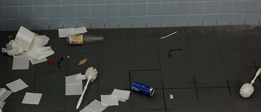 Ausschnitt: Glasflasche mit kleinem dunklem Fleck auf trockenem Fliesenboden, daneben trockene Handtücher und eine Toilettenbürste.