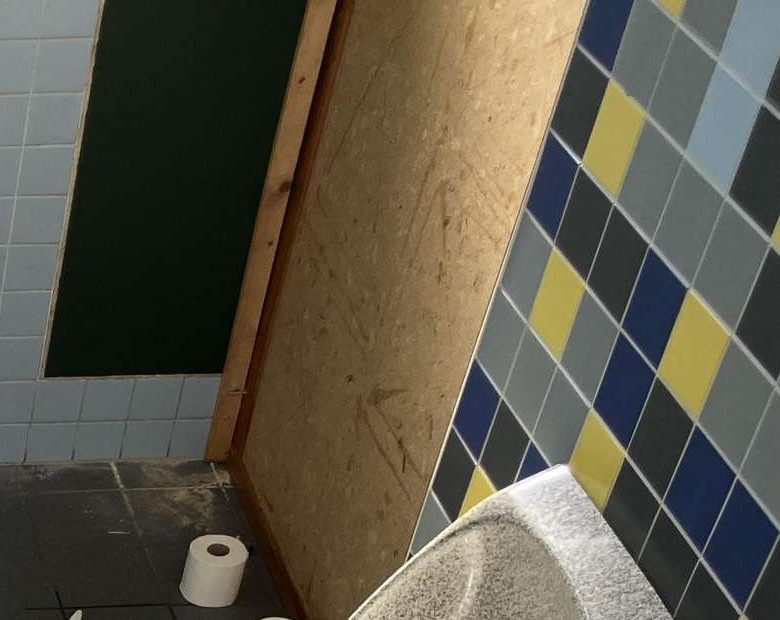 Ausschnitt: unverkleidete, zerkratzte Holzplatte als Wand neben den Mosaikfliesen.